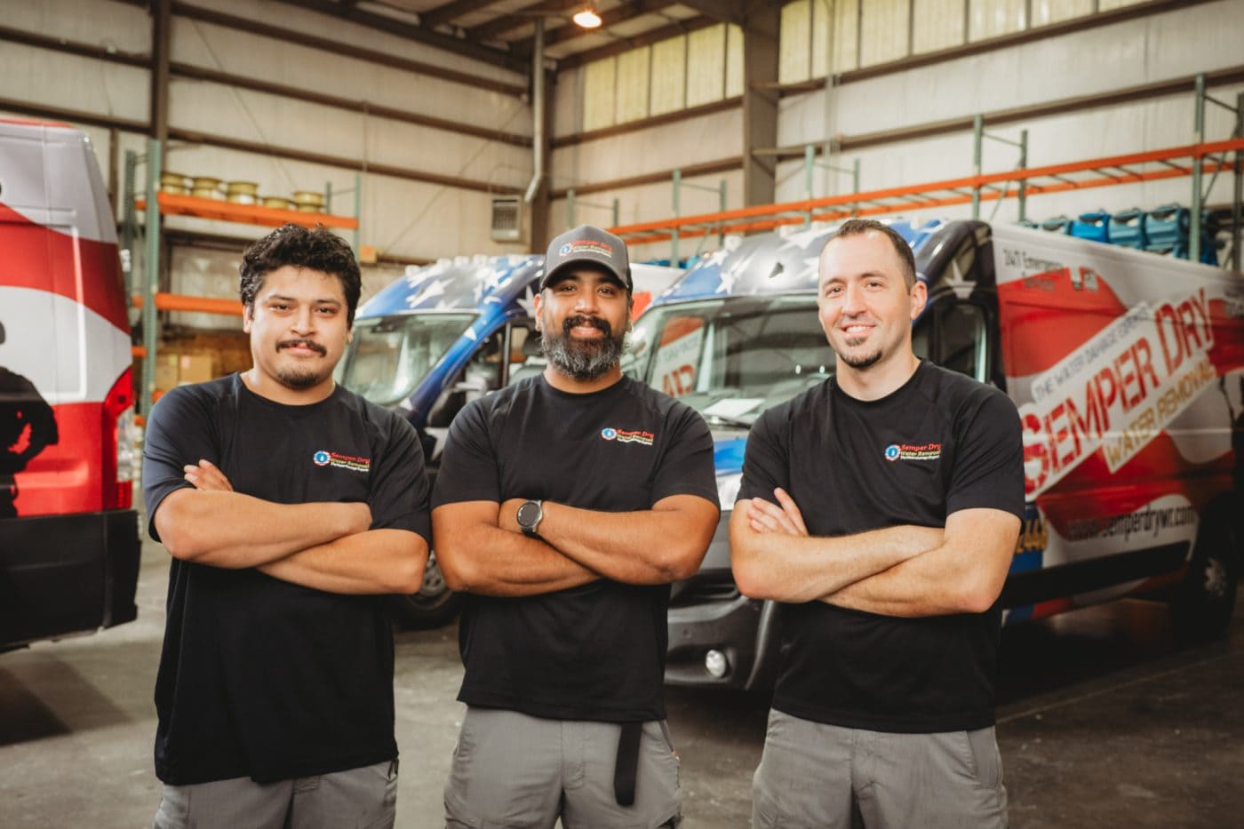 Three IICRC certified Semper Dry restoration technicians standing in front of patriotic wrapped service vans in the Virginia warehouse